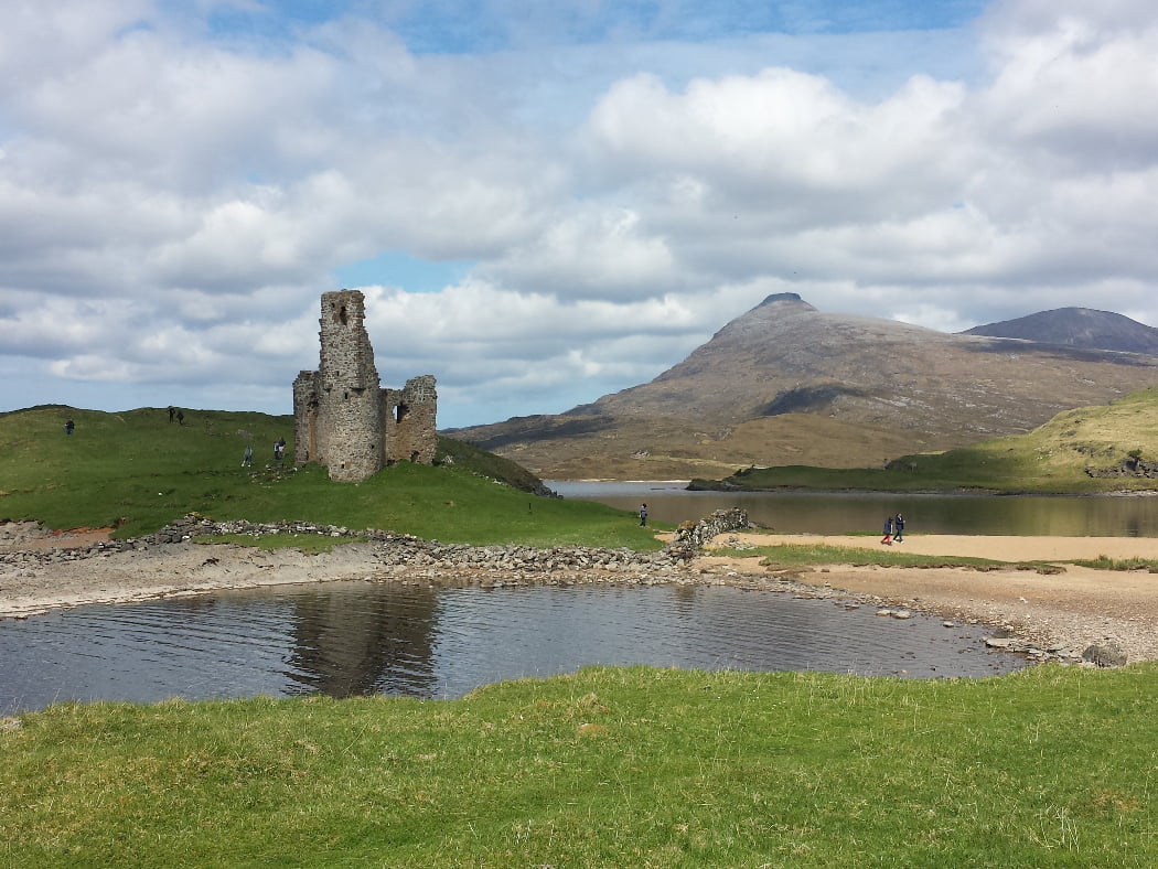 Just left of centre, the main focus of this picture is a ruined castle with a single ruined tower. It is built on a green hillock projecting into a lake. There are 9 or 10 people in the distance, walking around it. In the far right background a double-topped grey mountain rises towards a cloudy sky.
