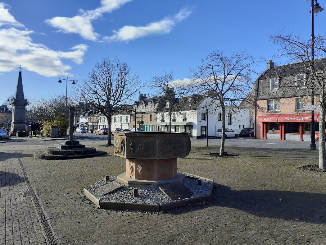 Extreme left stands the Clan Fraser of Lovat Memorial in Beauly visited on our tour from Inverness. Mid left stands the 1400's market pillar, and front and centre is a fountain that resembles a baptismal font. the urban scene of the square with the monuments is bounded to the right by the pretty three-storey buildings on the north side of Beauly Square. The sky is blue, and some small leafless trees stand down the centre of the square.
