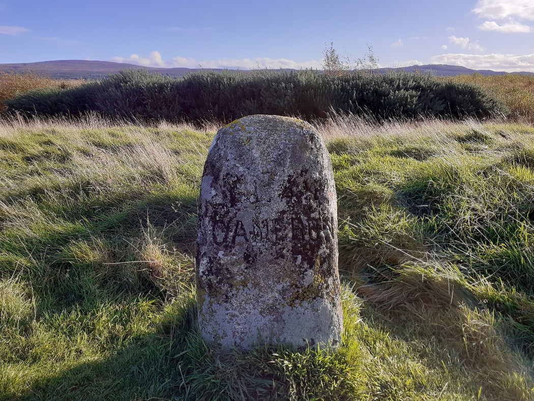 This is a close-up of a rough grave stone on Culloden Moor with the words Clan Cameron chiselled into it, visited on the Clan Cameron Tour from Inverness. The stone is in grass, with bushes behind, and a low and distant hill ridge in the background, under a blue sky.