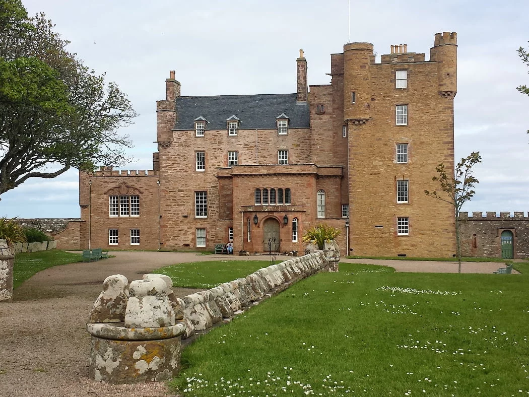 A red stone built castle of obviously differing ages. The 5 storey old keep with pepper-pot towers and flatter roof is at the right hand end. The middle section is 4 storeys and slate roofed, and the left hand end is two storeys and embattled. There are lawns with gravel drive in front, and a low wall running straight out of the picture towards us.