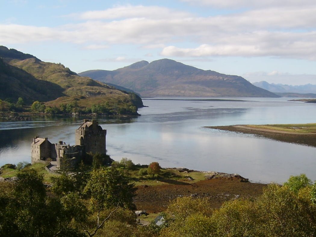 A castle, Eilean Donan, viewed from above on our Clan Mackenzie Tour from Inverness. It is Scottish Atlantic Seaboard design dating originally from 1200s. Behind is a blue arm of the sea, a wooded far shore. and in the distance middle and right are the jagged blue mountains of the Isle of Skye. The sky above is mixed blue and cloud.