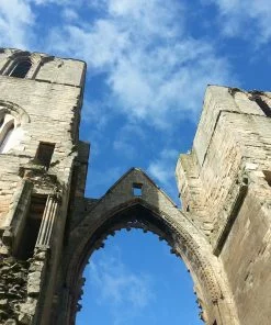 Two huge, ruined, medieval, flat-topped, cathedral towers in Elgin Cathedral on the Local Dark Deeds Tour. The sky above is quite a strong blue with wisps of white cloud. The stone is predominantly a red sandstone. A ruined wall ties these two towers together, and holds the ruined framework which would originally have held a massive arched window (the largest in the cathedral).