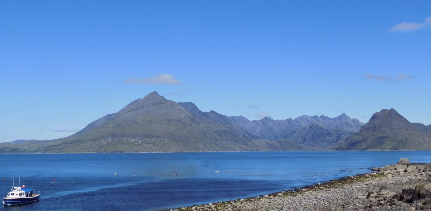 Our Isle of Skye Tours from Inverness can include a boat trip. The foreground is an ocean strait with the boat at sea to the left. In the background, across the water, is the long Jagged ridge of the Black Cuillin Mountains. The sky is blue and nearly cloudless.