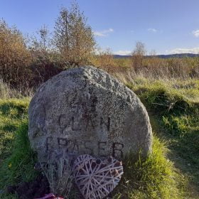 Foreground is the Fraser Clan Grave Stone on our Clan Fraser of Lovat Tour from Inverness. It is about 0.5m in height, grey with a rounded top, and the words Clan Fraser carved into the front. A woven heart ornament lies on the ground in front of the stone. There are shrubs and trees behind, and a bare hilltop ridge in the far distance. Above, the sky is blue.