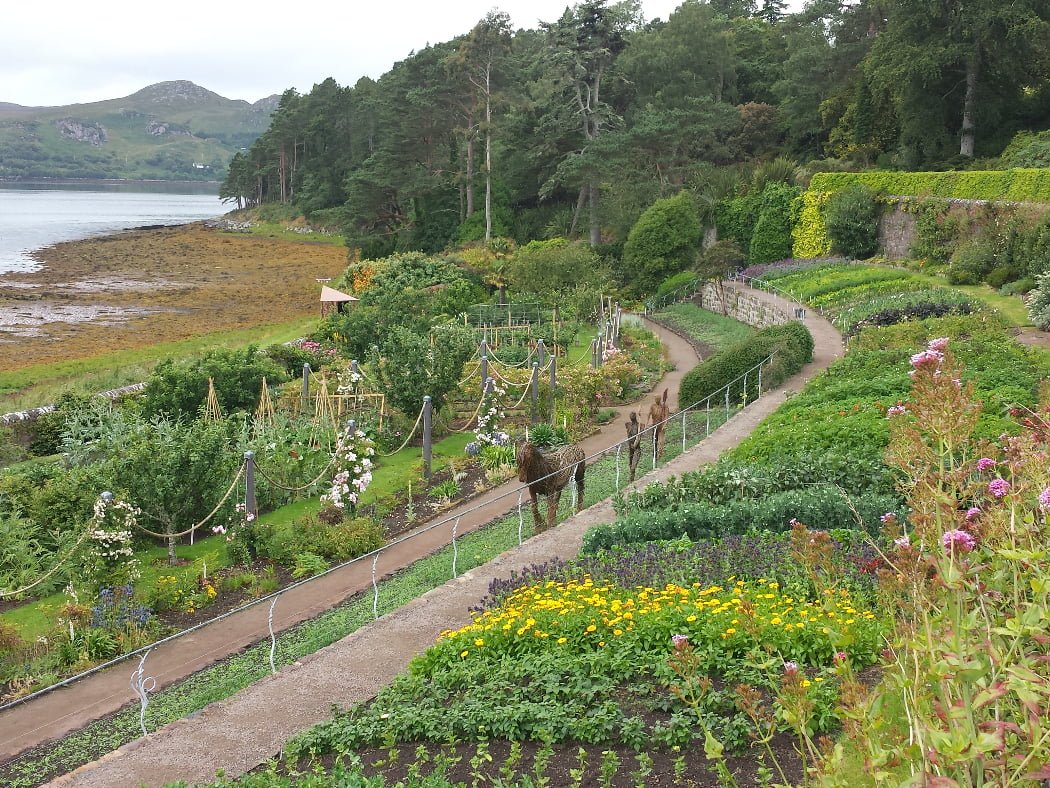 Colourful Victorian walled garden terminating to the right in a retaining wall against the wooded hillside, while to the left is the seashore. Farther left still is the distant rugged hills on the far side of the sea inlet. Along the main path in the gardens is a wicker sculpture consisting of a horse clopping towards us, followed by two wicker figures walking behind with shovels over their left shoulders.