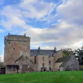 Kilravock Castle, near Inverness, is on our tour because it is the seat of Clan Rose. The foreground is grass. There are trees to left and right. A blue sky with clouds is above. The castle fills most of the picture. Left hand half of the castle is a tall square medieval grey stone keep. The right half is newer with portions from the 18th and 19th centuries.