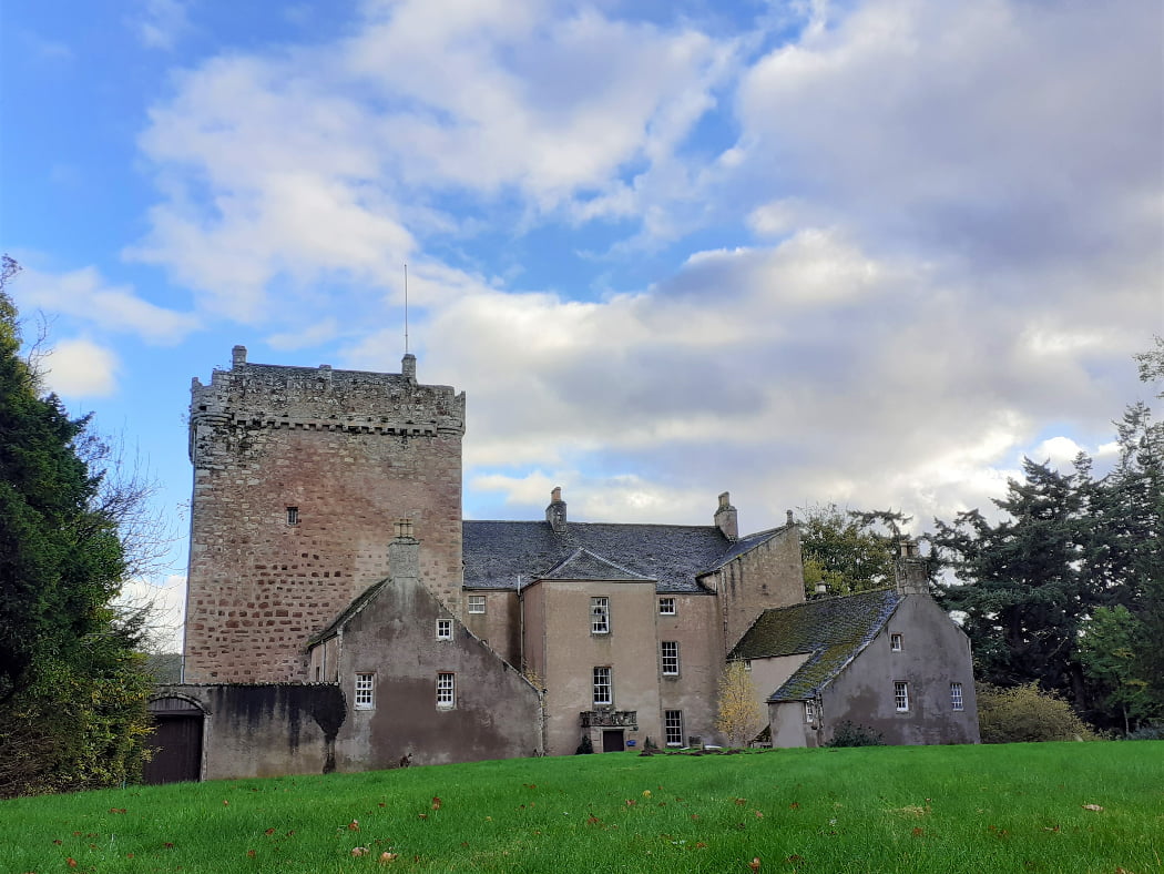 Kilravock Castle, near Inverness, is on our tour because it is the seat of Clan Rose. The foreground is grass. There are trees to left and right. A blue sky with clouds is above. The castle fills most of the picture. Left hand half of the castle is a tall square medieval grey stone keep. The right half is newer with portions from the 18th and 19th centuries.