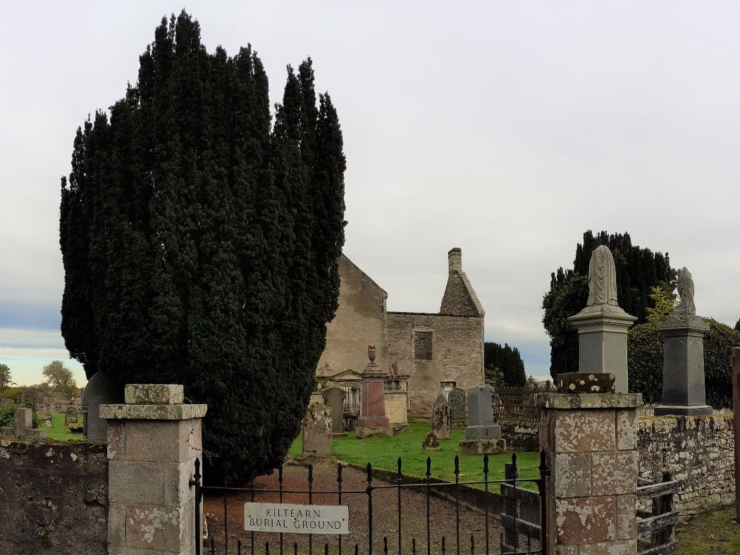 The foreground is a churchyard wall, and pillars flanking a black iron gate. Behind the left pillar is a large shaped yew tree, and appearing behind and to the right of it the roofless ruin of a stone church. There are many tombstones of various sorts to be seen standing in the grass, in the churchyard. The sky is white.
