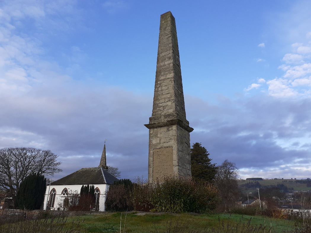 An old stone obelisk on a slight grassy rise projects into a blue sky. On the front is a large brown-coloured rectangular plaque which is too far away to be read. low shrubs surround the base. there is a white church with grey stone steeple some distance behind the obelisk to the left. to the right, behind the obelisk is a distant hill, with farmland and trees. Closer to the right, streetlights hint that this is an urban location.Mackenzie Monument - Clan Mackenzie Tour from Inverness