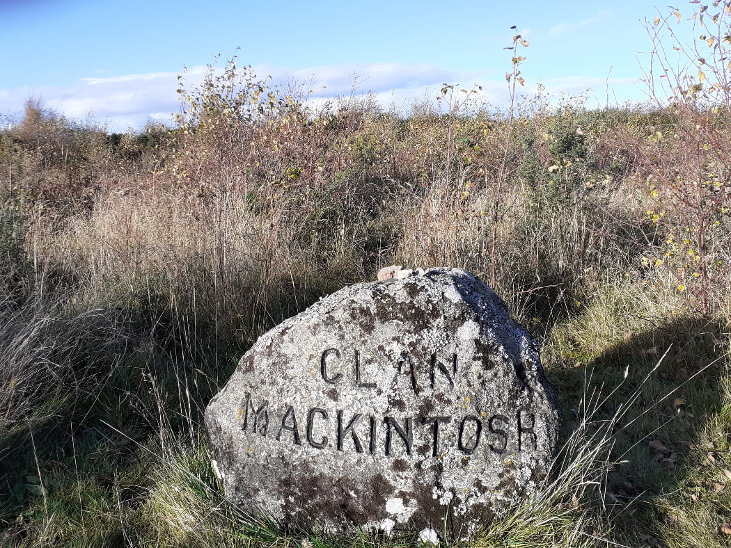 This is a close-up of a rough grave stone on Culloden Moor with the words Clan Mackintosh chiselled into it, visited on the Clan Mackintosh Tour from Inverness. The stone is in grass, with bushes behind, under a blue sky.