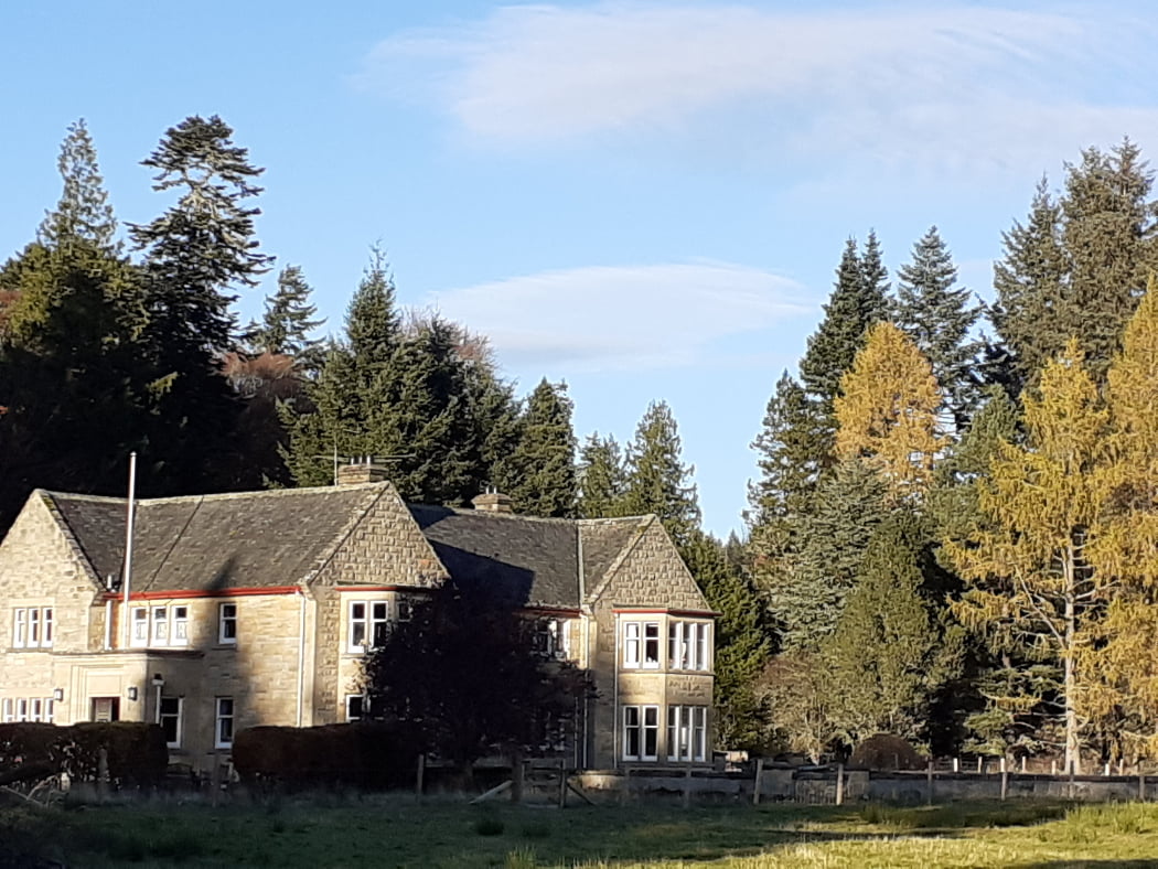 Moy Hall, near Inverness, visited on the Clan Mackintosh Tour is from the centre to the left. It is a 2-storey stone-built 20th century building with bow windows to the front and a grey slate roof. There are large mature trees behind, and a blue sky above.