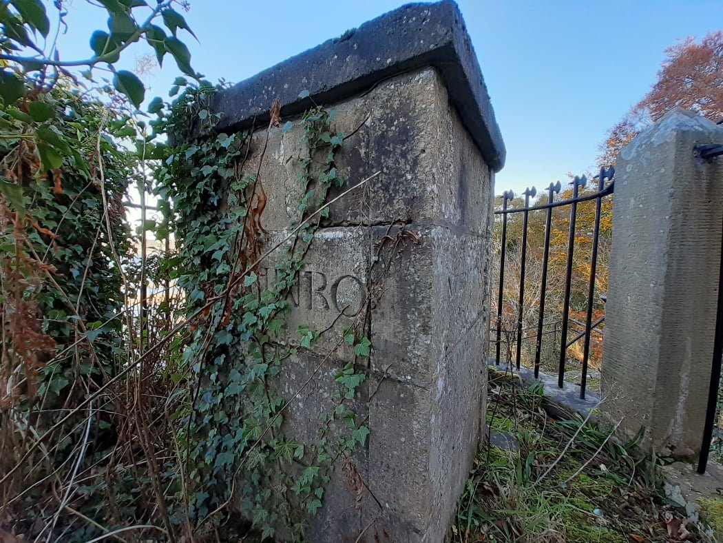 The bulk of the picture is a square grey flat-topped stone monument (about 4 feet high) covered in ivy. Through the Ivy the letters NRO are just visible. The MU at the beginning of the word is under the vegetation. To the right are the iron railings which surround the site. A blue sky above.
