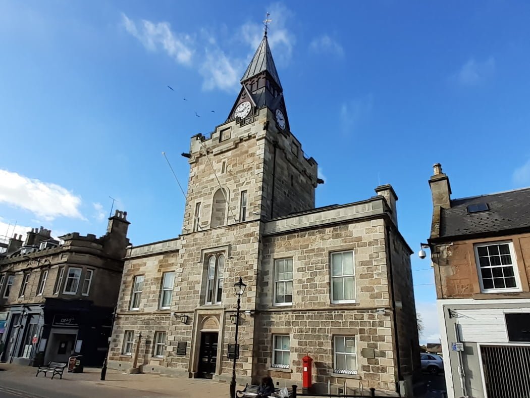 A civic building dating from the mid-1800's, whose main block is 5 windows wide and 2 storeys high. A large black door is centrally placed on the frontage and above the whole edifice rises an embattled tower with spire with clock faces. The sky above is blue.