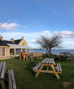 The yellow-coloured, slate-roofed Storehouse of Foulis restaurant and Clan Munro exhibition is to the left in the picture, on the tour from Inverness. To the right is an inlet of the blue sea. The sky is blue with a few white clouds. The foreground is all grass and 8 or 9 wooden picnic table sets.