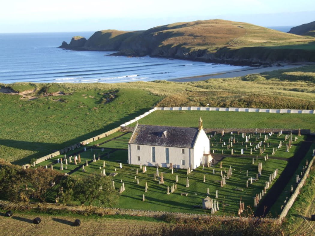 Arial shot of a two storey church, light grey with a dark grey slate roof. It stands in a square churchyard with many tombstones around it. There is a cove with a beach behind the church, and beyond it a bare grassy headland pushing out into the fairly calm blue ocean.
