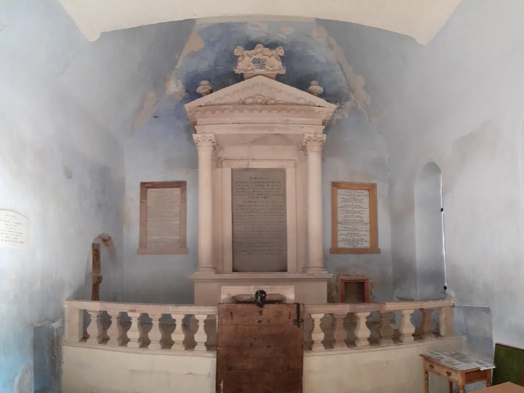 We are inside a light-blue painted mausoleum. Directly in front of us is a monument in white marble occupying the whole end wall. There is a platform surrounded by a balustrade, and centrally on the platform is an edifice comprising pillars to either side of a portico surmounted by an ornate pediment. There is a large tablet with writing on the wall at the rear of the portico, and a smaller tablet to each side.