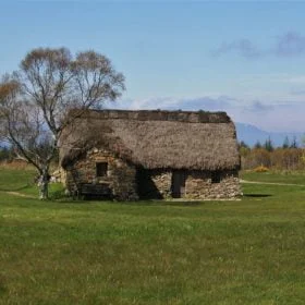 There is grass in the foreground, and most of the image is a stone-built one-storey cottage with a heather thatched roof that we visit on our Multi Day Outlander Tours from Inverness. There is a deciduous tree to the left, and behind the cottage is a mountain in the far distance. On the horizon are more trees, and the sky above is blue.