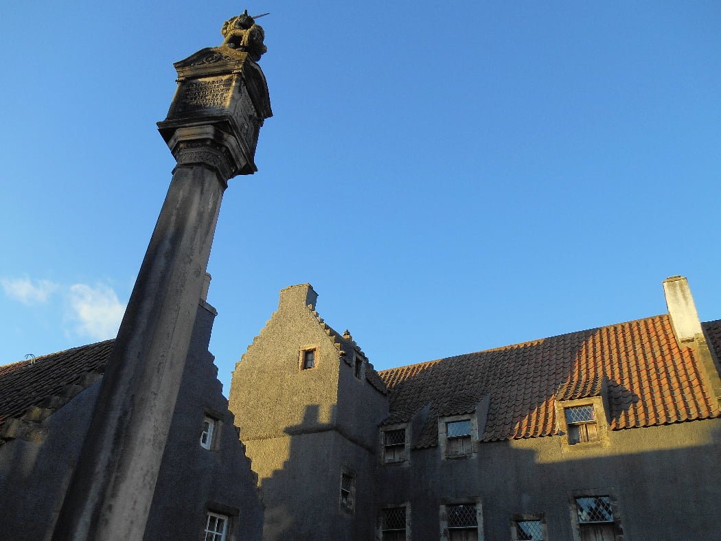 This is a close-up of the Culross (Cranesmuir) market pillar on our One Day Outlander Tour from Glasgow. It is surmounted by a unicorn, and the sky above it is cloudless blue. Two buildings are visible, one to the left, behind the pillar, and the other, straight ahead, is Geilis Duncan's house from the filming. The buildings are painted dark grey in preparation for the filming.