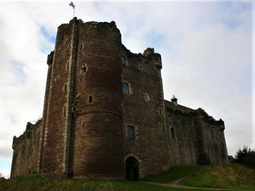 The great gatehouse of Doune Castle (Castle Leoch) rears into a cloudy but blue sky, on the One Day Outlander Tour from Edinburgh. There is a flagpole on the battlements of the tower, flying the Scottish flag.