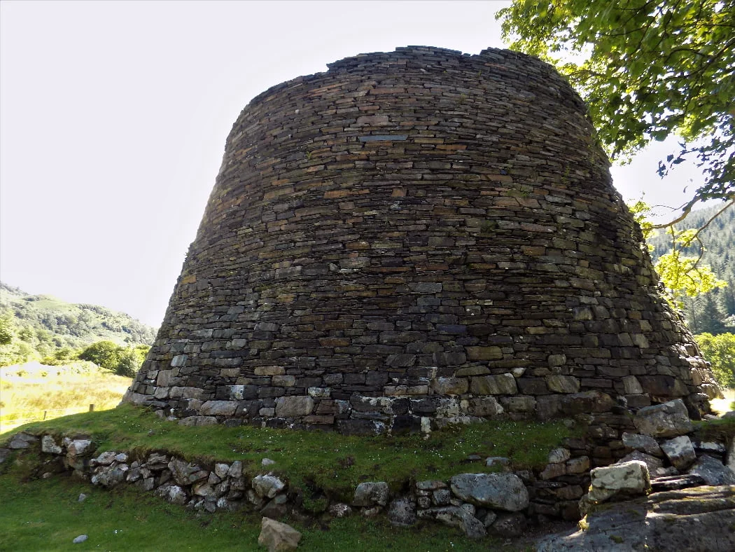 The 30-foot ruined tower filling this image is a broch. A large deciduous tree is to the right, and in the distance, to the left is a hillside covered in deciduous trees (all in leaf). It is a sunny day, though we are in the shade of the tower, and the sky is a cloudless very pale blue. and there is a blue sky above.