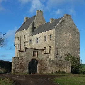 On our One Day Outlander Tour from Edinburgh is an ornate partial wall with archway, behind which the four-storey grey stone Towerhouse of Midhope Castle (Lallybroch) stands. The sky above is blue.