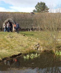There is a small pond in the foreground of this shot taken on our Multi Day Outlander Tours from Edinburgh. Up the grass slope behind it is a long thatched building with a single door towards the left hand end. There is a man and a woman dressed in farming 1700's costume at the left hand end of the building and five visitors in front of the door. The sky above is cloudy.