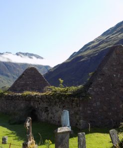 The churchyard with tombstones in the foreground is visited on our Multi Day Outlander Tours from Glasgow. This is a roofless ruined church filling most of the image. Behind, rising into a pale blue sky, are some very rugged mountains with just a little tonsure of clouds on them.