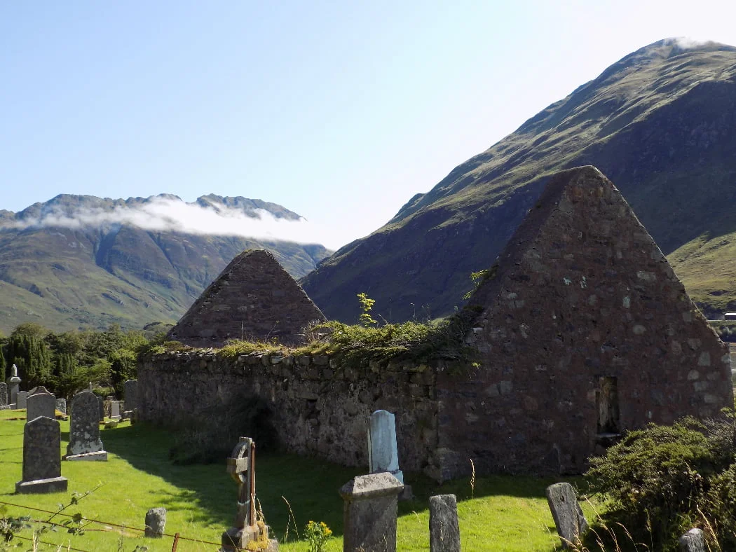 The churchyard with tombstones in the foreground is visited on our Multi Day Outlander Tours from Glasgow. This is a roofless ruined church filling most of the image. Behind, rising into a pale blue sky, are some very rugged mountains with just a little tonsure of clouds on them.