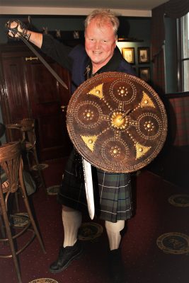 Hugh Allison is in the foreground in this image. He is inside a public room, and is kilted. He is holding a sword overarm in his right hand, and a shield and dirk (dagger) in his left hand. He is lunging towards the camera.