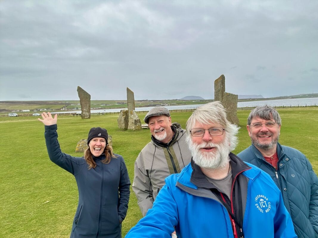 Four people three men and one woman, standing facing a camera in a field of green grass, with a circle of standing stones to rear, it is a overcast day, the woman is waving the men are smiling