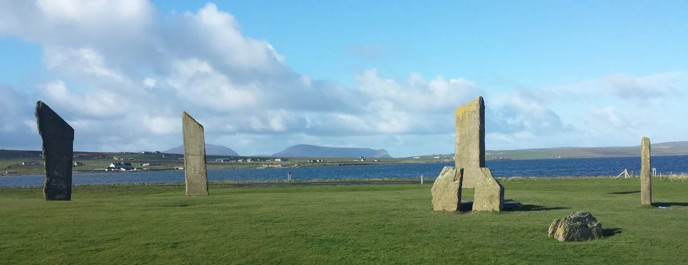 wide image of seven of the Standing Stones of Stenness on Orkney. Behind is the Loch of Stenness, lots of distant farms and farmland and in the far background the high hills of the island of Hoy.