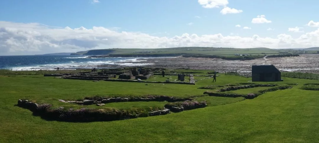 A sunny but windy/wild looking day on the tidal island called the Brough of Birsay. In the green grass of the foreground we can see the low foundations of Pictish and Viking settlement. In the middle distance we see the tidal crossing and in the background are the low rising green hills of Orkney Mainland.