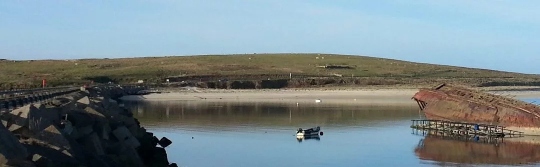 This is a wide picture of the Churchill Barriers we visit on our Orkney Tours from Inverness. On the left is the concrete block built WWII causeway called a Churchill Barrier. On the right is the rusting hulk of a war-time blockship. The ocean is still, and in the background a gentle green hill rises above a sandy beach.